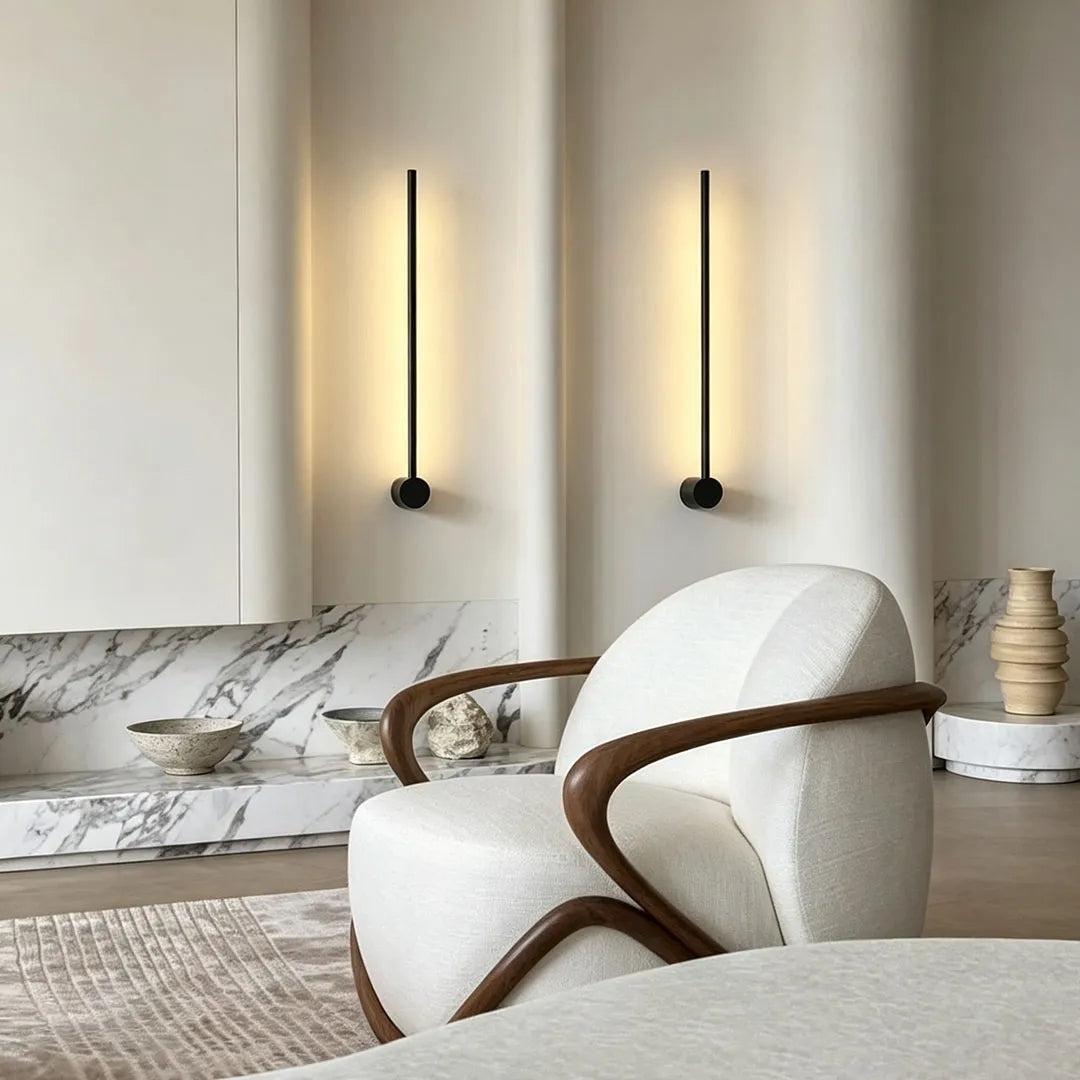 Linear LED wall lights mounted in a minimalist interior corner beside a sculptural lounge chair and marble bench.
