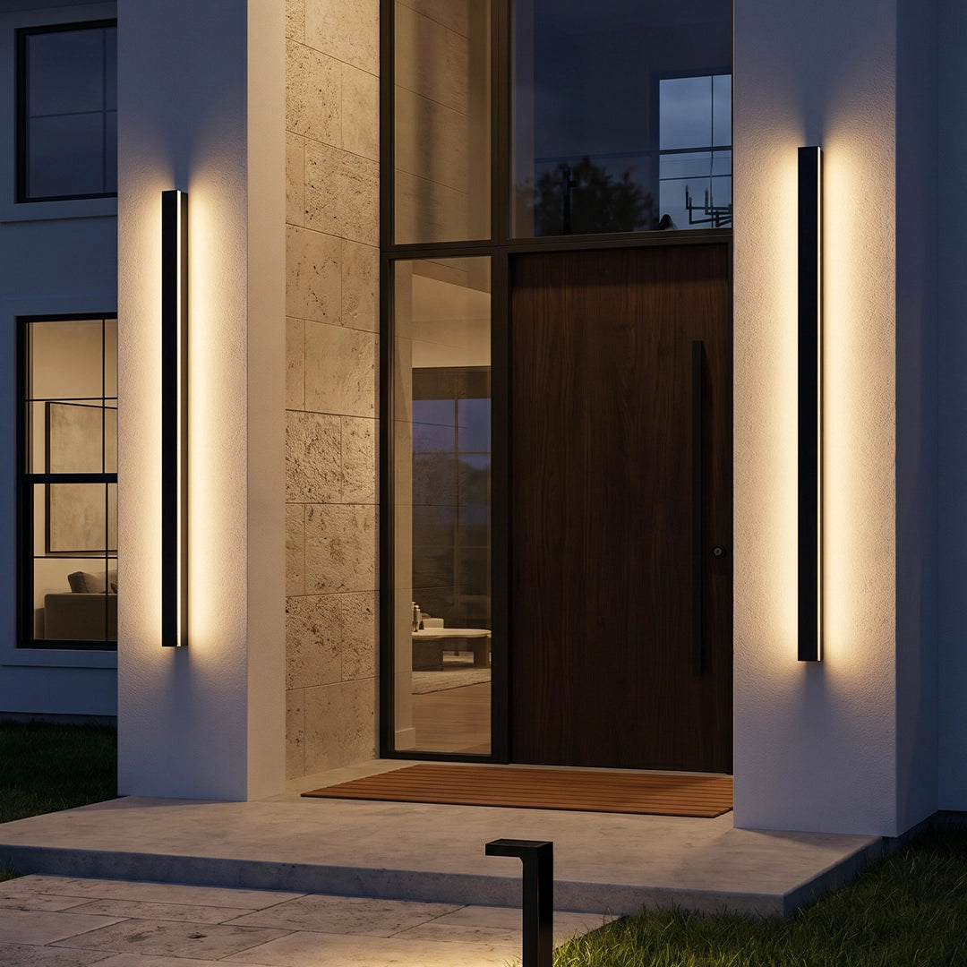 Linear outdoor wall sconce mounted beside a modern front door on a tall stone entry wall at dusk.