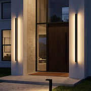 Linear outdoor wall sconce mounted beside a modern front door on a tall stone entry wall at dusk.