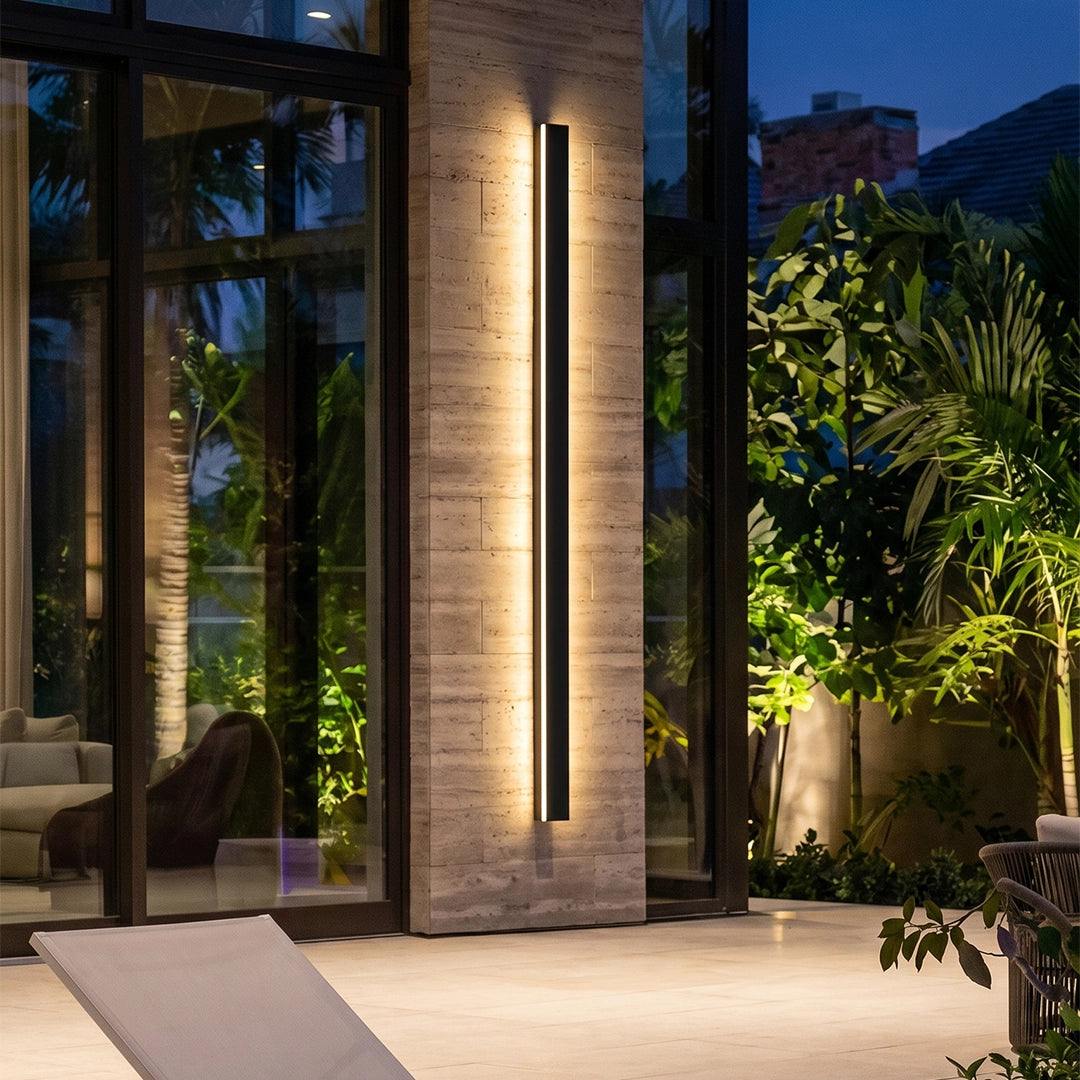 Linear outdoor wall sconce mounted on a stone patio wall beside tall glass doors and lush plants.