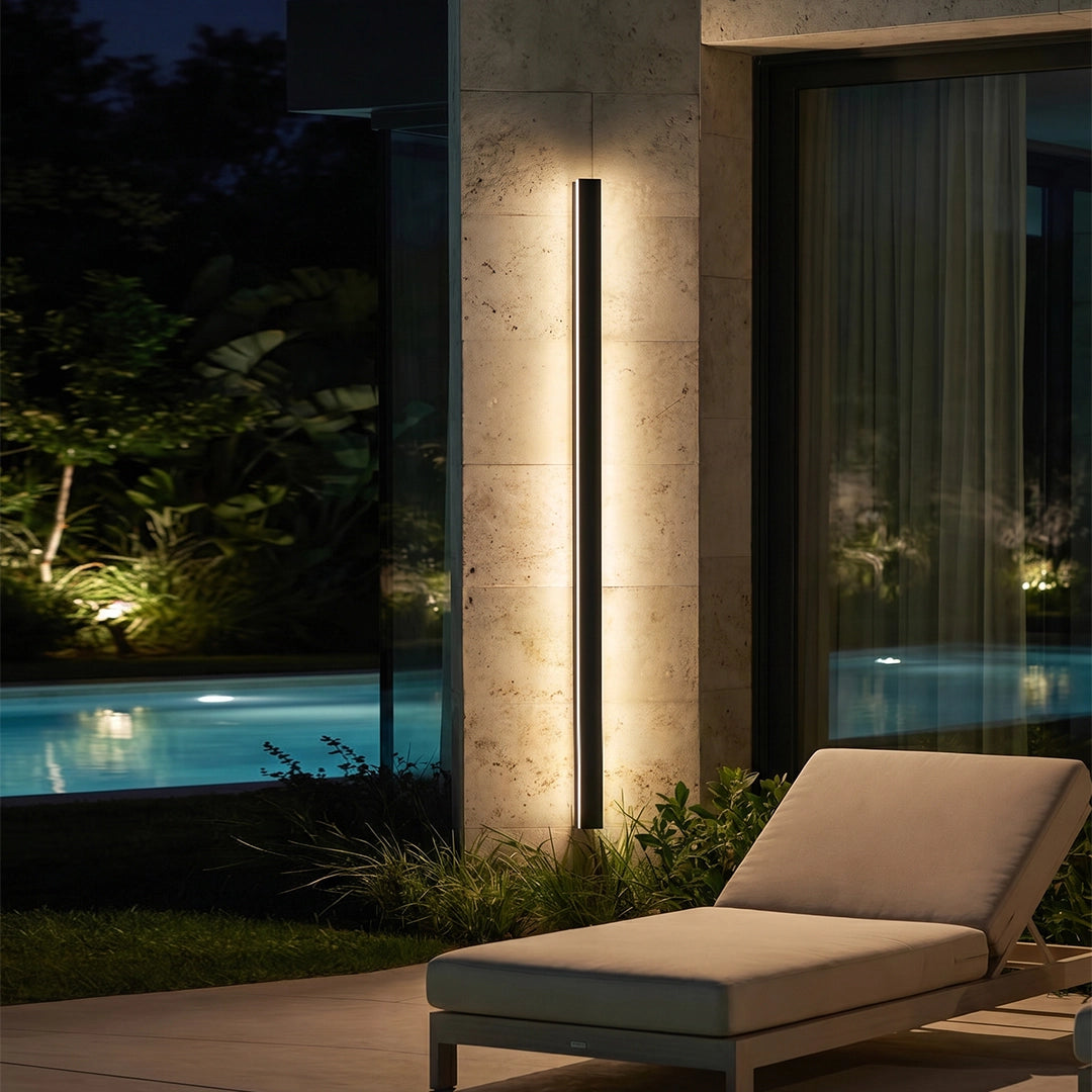 Linear outdoor wall sconce mounted on a stone wall beside a pool and lounge chair at night.