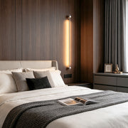 Modern long linear wall sconce installed beside bed with warm vertical LED ambient lighting.
