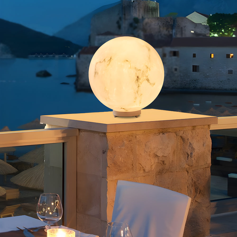 Outdoor pillar lights with a marble finish, casting a warm glow on a stone pillar by the waterfront.