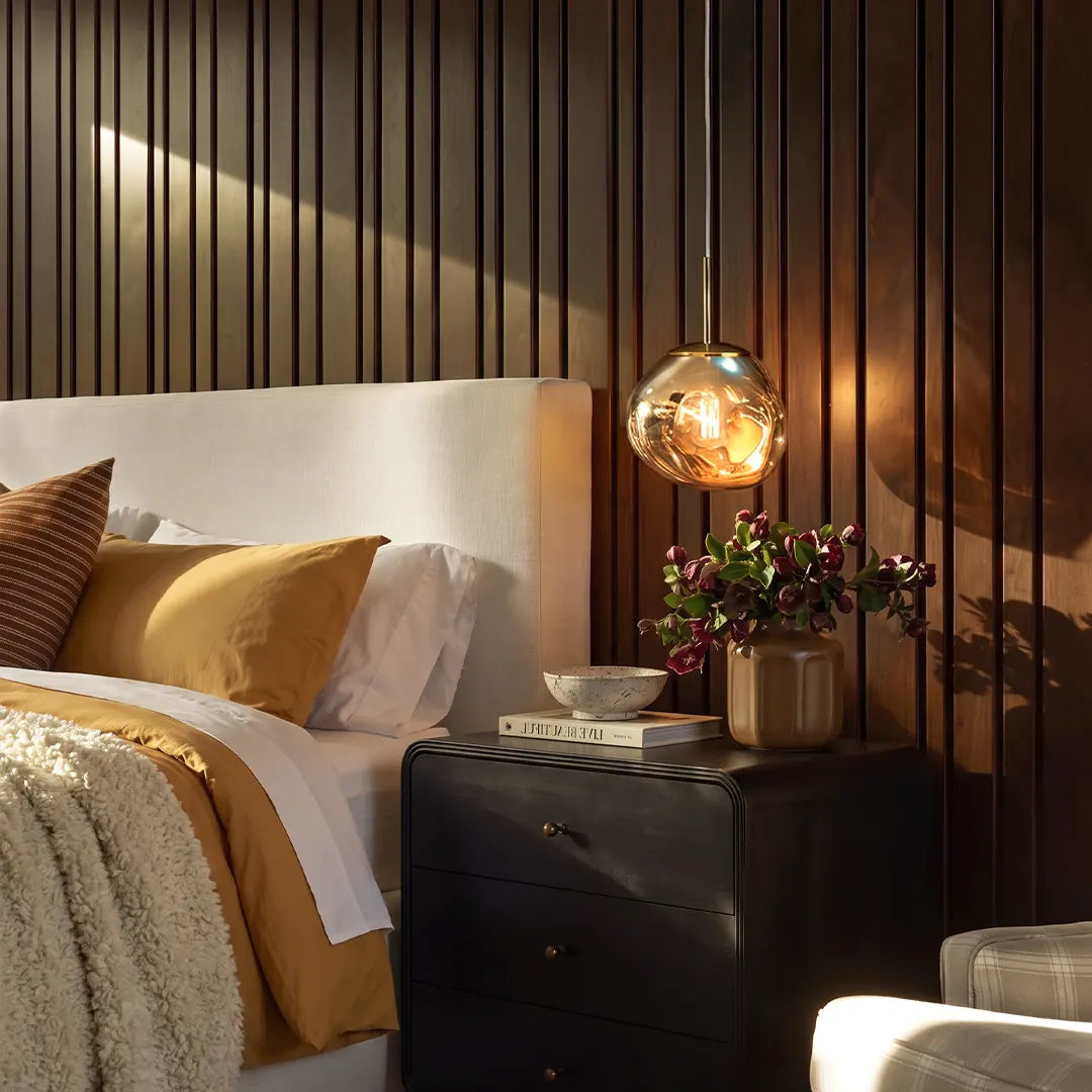 Melt pendant light with a reflective amber globe shade hanging beside a bed above a black nightstand in a warm modern bedroom.