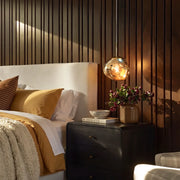 Melt pendant light with a reflective amber globe shade hanging beside a bed above a black nightstand in a warm modern bedroom.