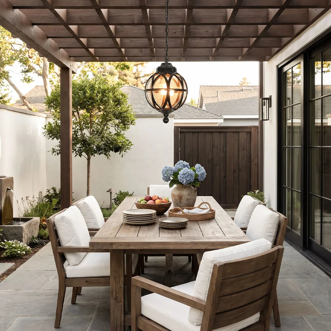 Metal orb pendant light suspended above an outdoor dining table under a pergola in a contemporary courtyard.