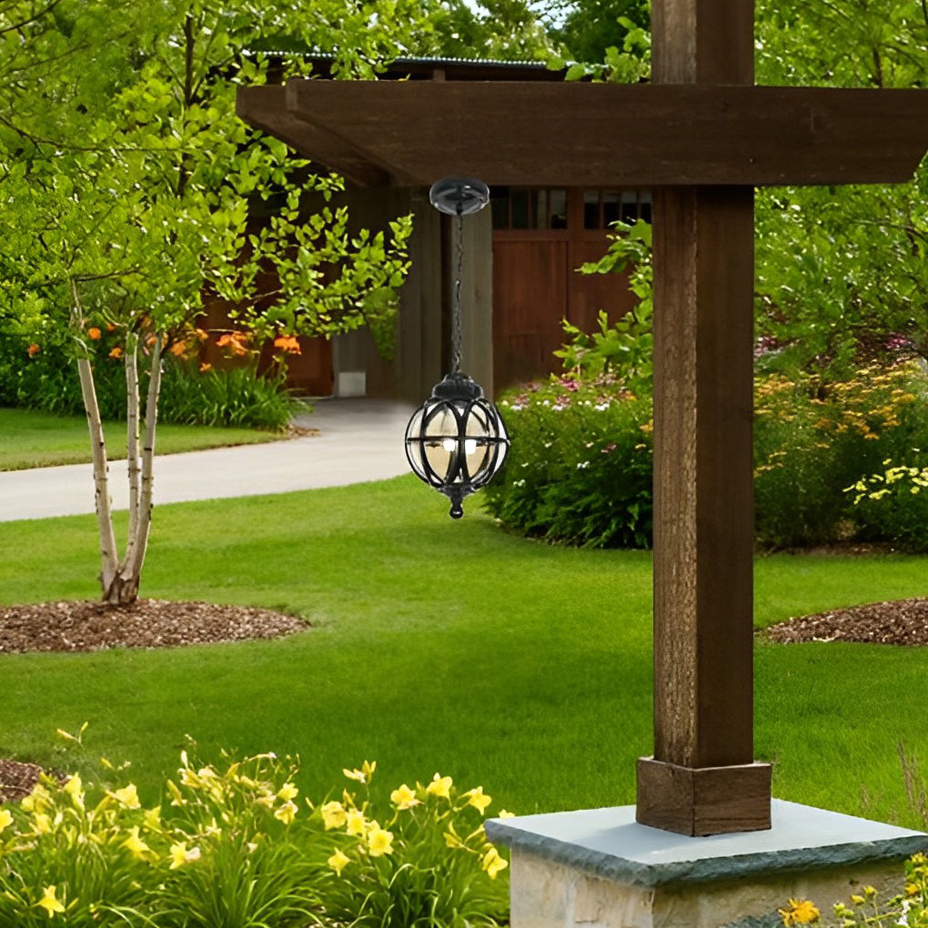 Metal orb pendant light mounted under a wooden outdoor structure, illuminating a landscaped yard area.