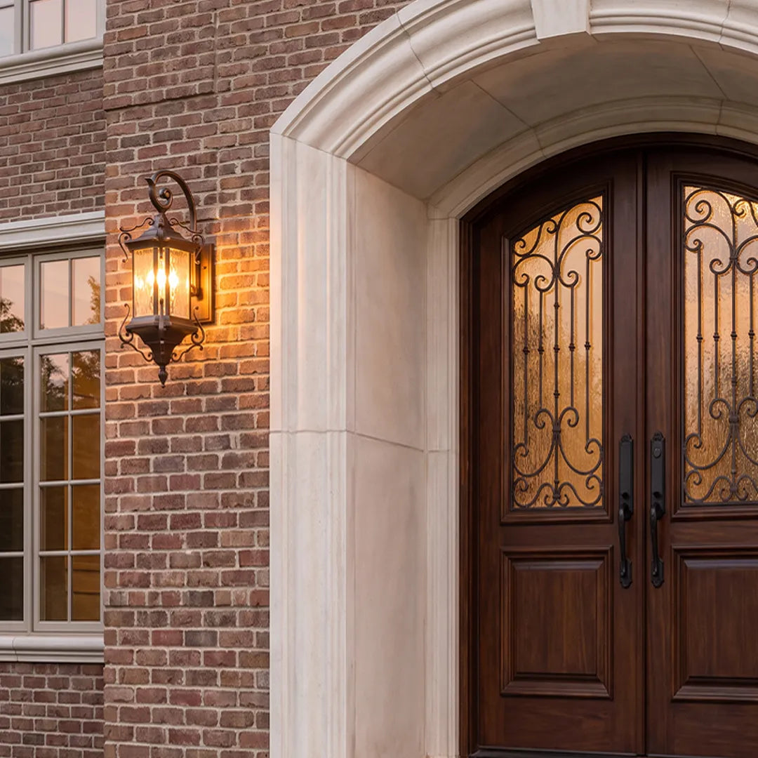 Metal outdoor wall sconce beside double wood entry doors, showing a decorative lantern design with warm exterior illumination.