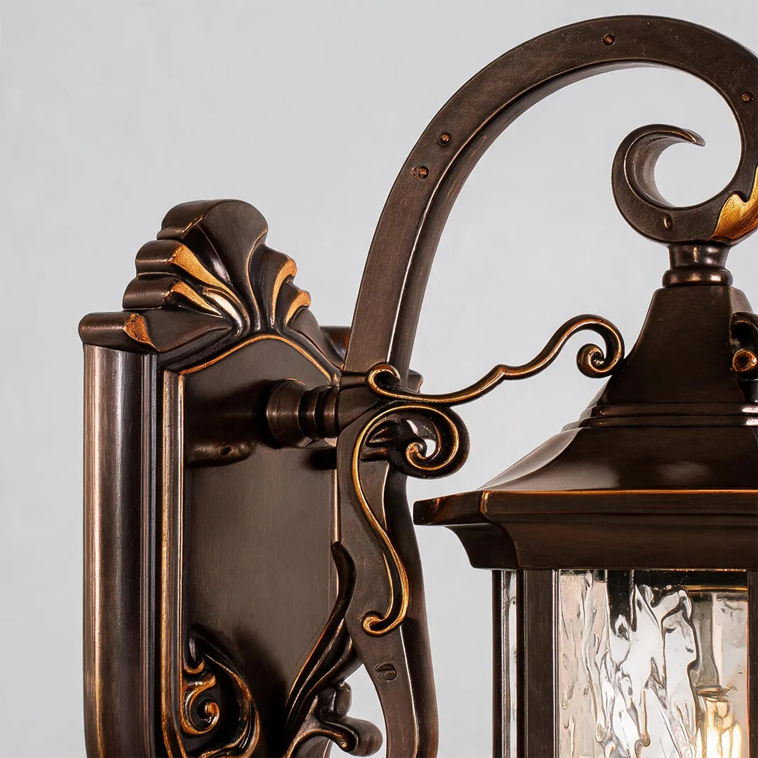 Metal outdoor wall sconce close-up showing the ornate backplate, scroll arm, and lantern top detail in a bronze finish.