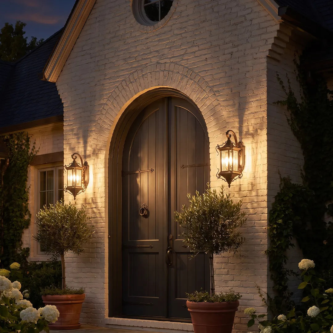 Metal outdoor wall sconces mounted beside a dark arched front door, adding warm lantern-style lighting to a white brick exterior.