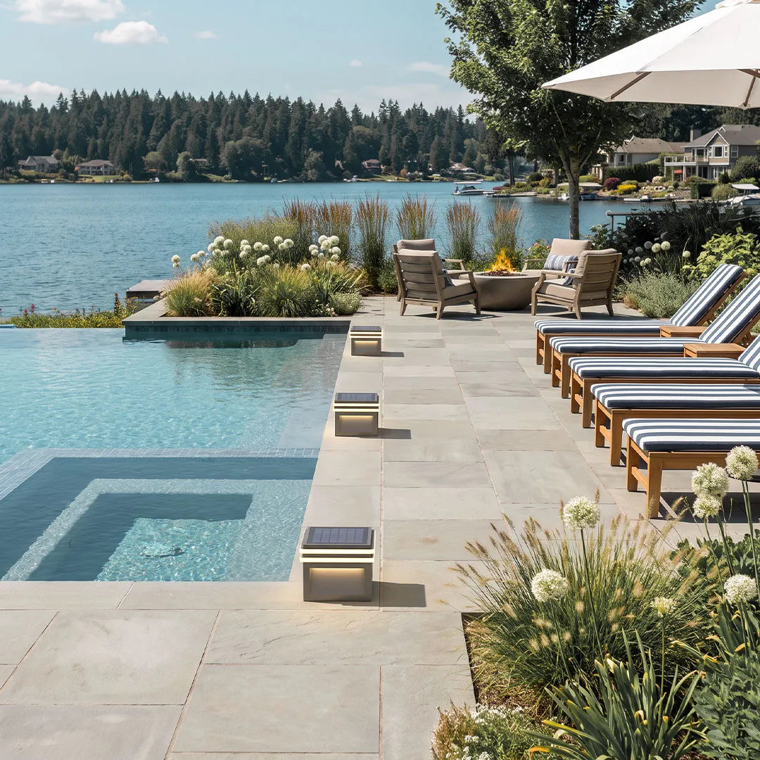 Metal pillar lights lining a poolside walkway beside a lakefront patio and lounge chairs.