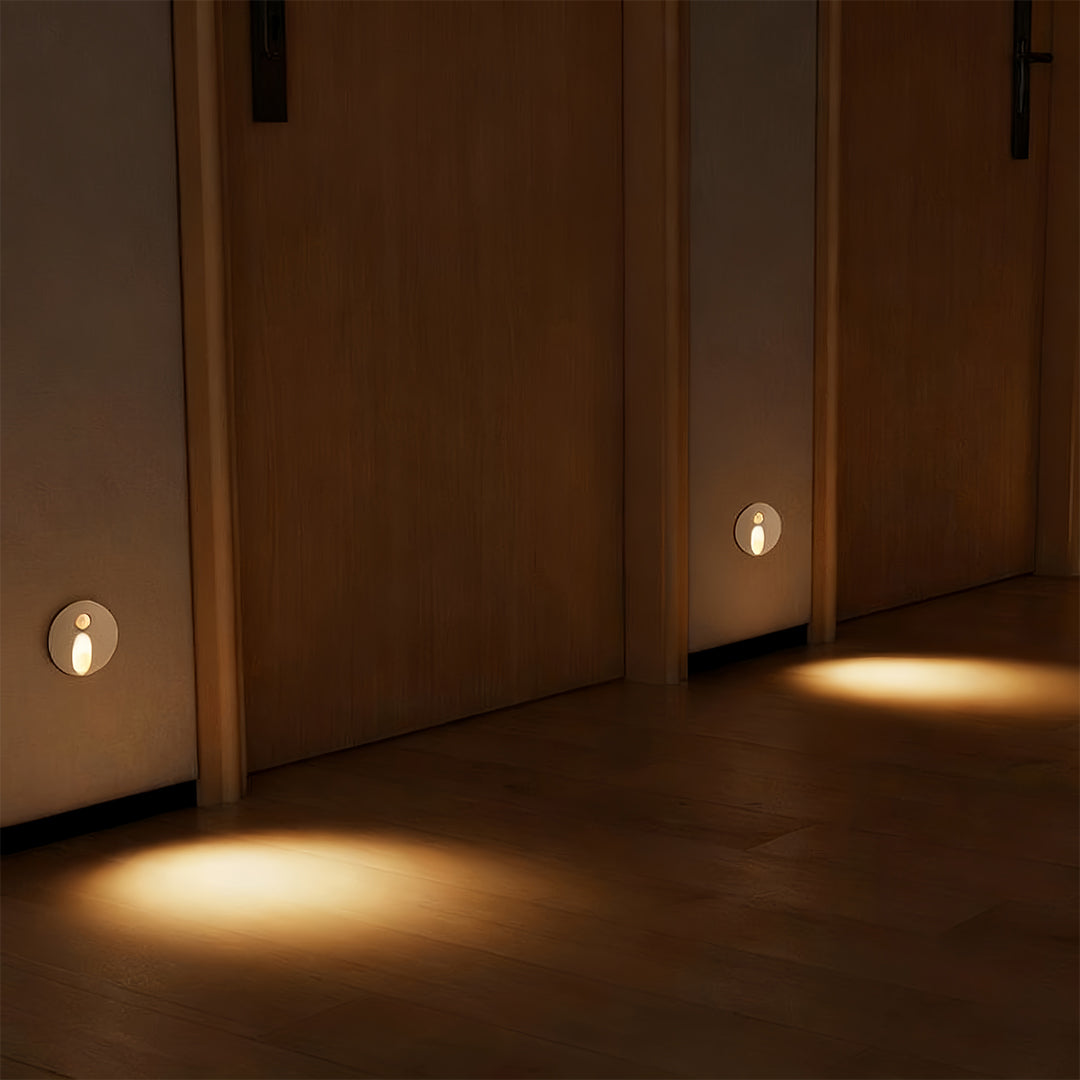Minimalist interior stair step lights placed near the baseboard in a hallway, casting a wide, inviting spotlight onto the dark wooden floor near a doorway.
