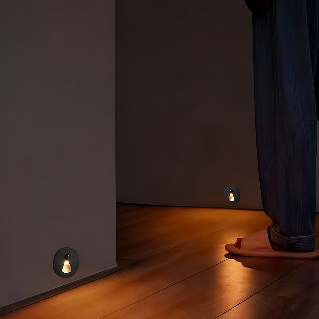 Minimalist interior stair step lights positioned low to the ground on a dark wall, casting a subtle, focused glow onto the wooden floor next to a flowing curtain.