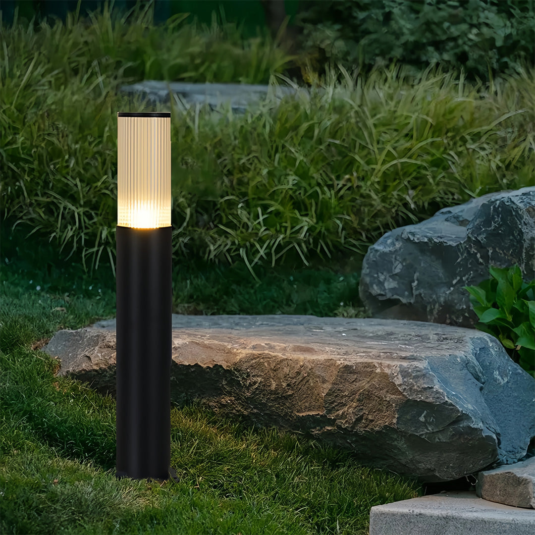Modern black bollard path light with a fluted glass top illuminating a landscaped garden bed.