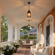 Modern black outdoor pendant light hanging above a covered porch seating area with white ceiling panels and brick accents.