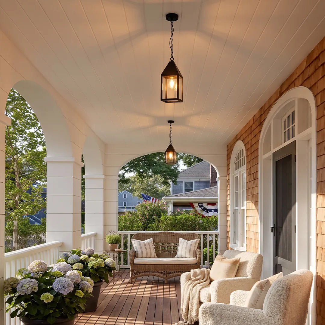 Modern black outdoor pendant light hanging above a covered porch seating area with white ceiling panels and brick accents.