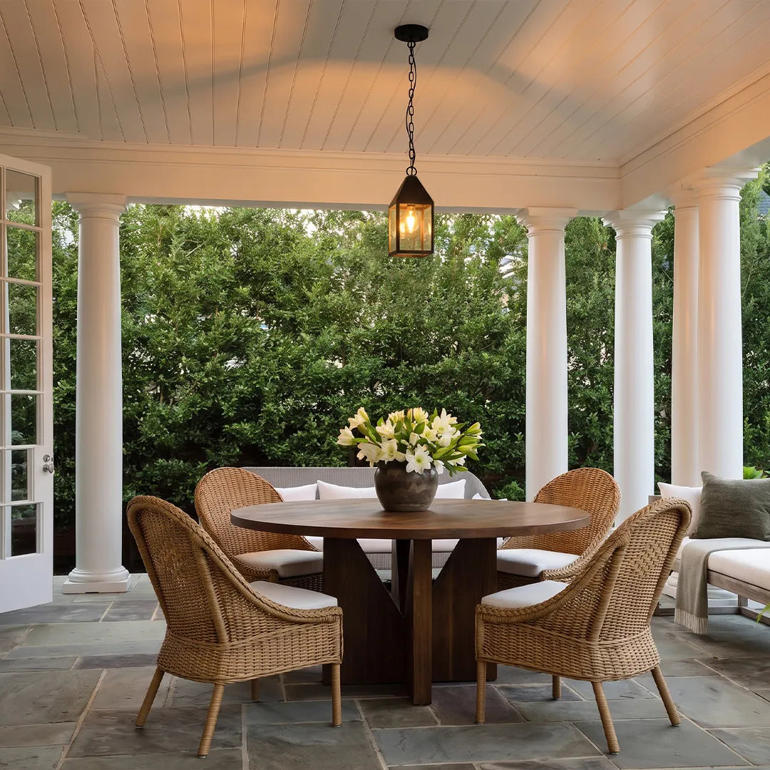 Modern black outdoor pendant light suspended over a round dining table on a bright covered patio with wicker chairs.