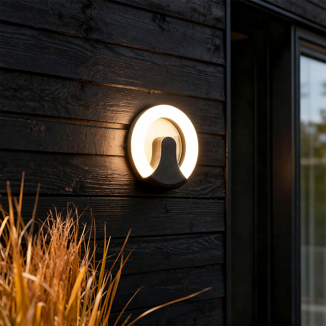Modern black wall sconce light featuring glass shade chandelier styling creating warm ambient glow on dark wood exterior wall.