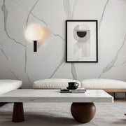 Modern black and white globe wall light mounted on a marble-textured wall above a low seating area in a minimalist room.