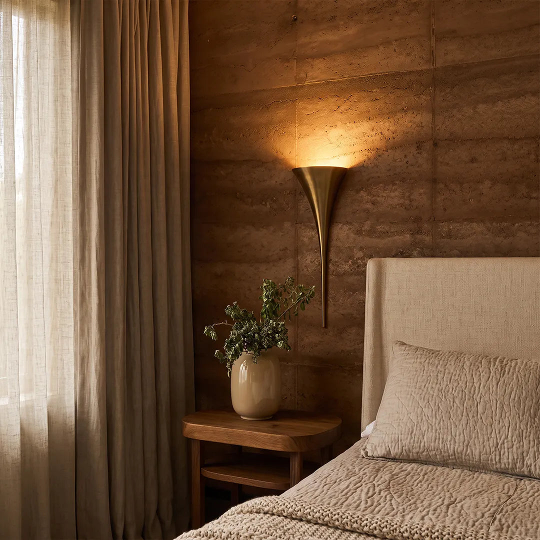 Modern brass wall sconce casting upward light beside a bed in a warm wood-accent bedroom.