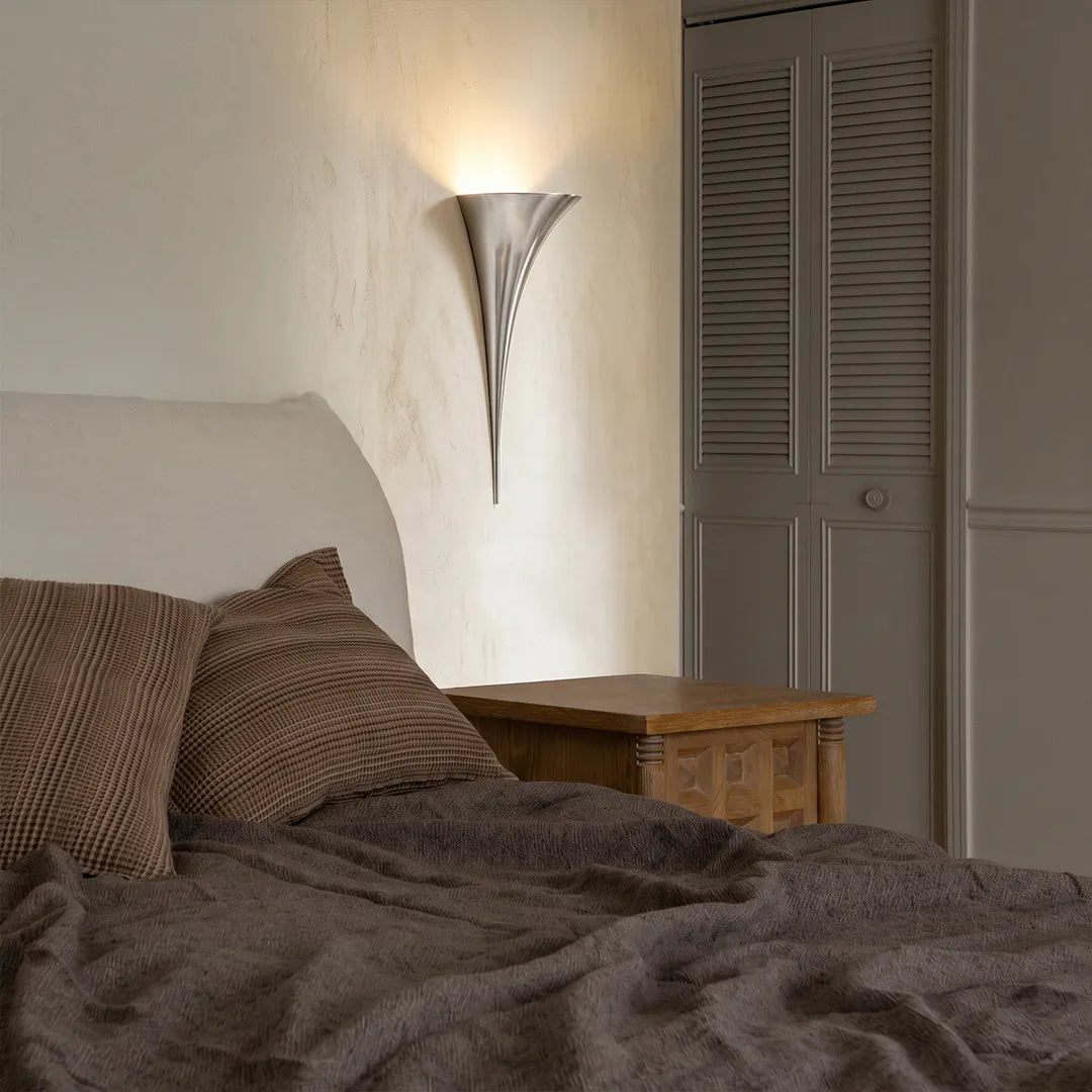 Modern brass wall sconce in a chrome finish mounted above a bedside table in a minimalist bedroom.