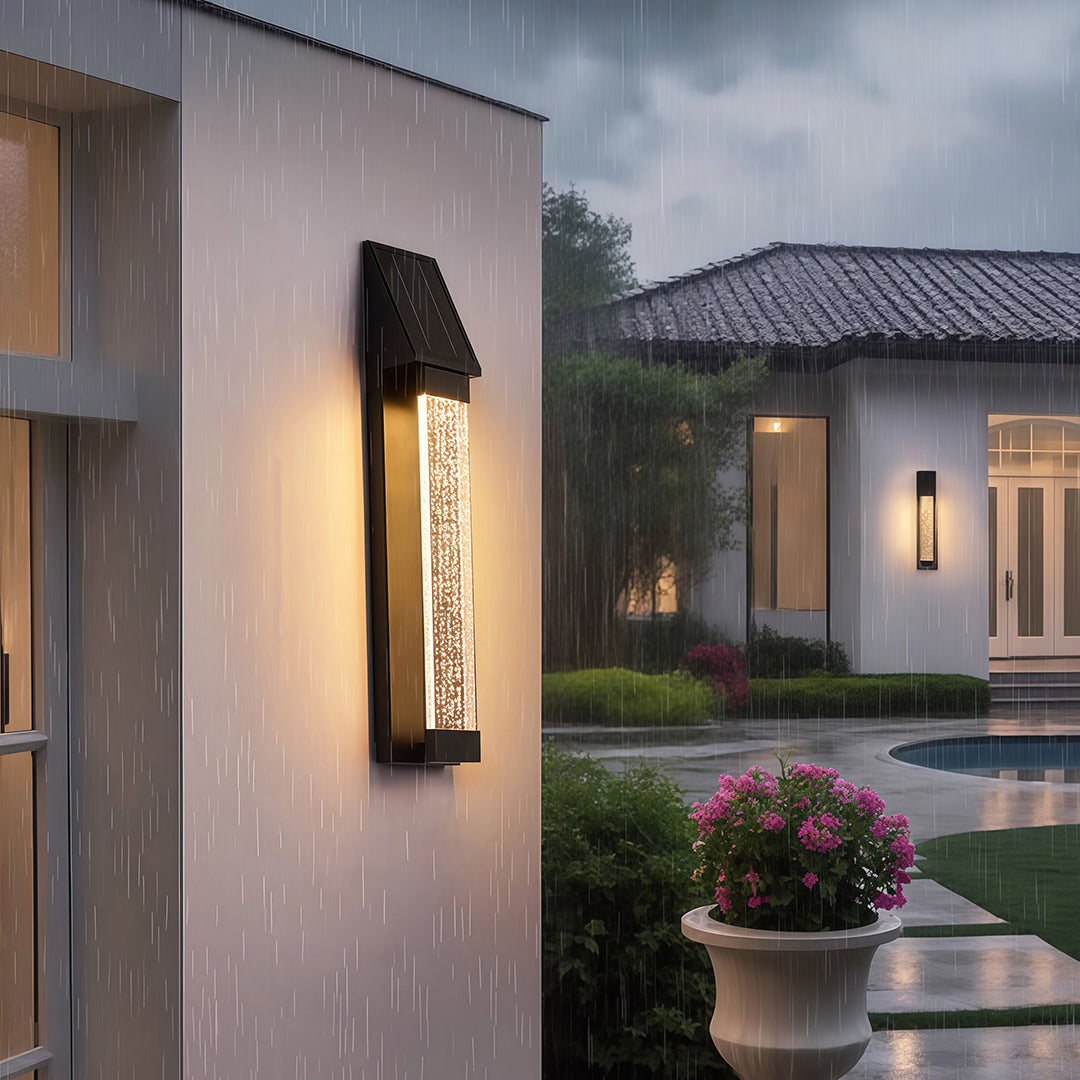 Black outdoor lighting sconce with a textured glass front, mounted beside a modern gate entrance.