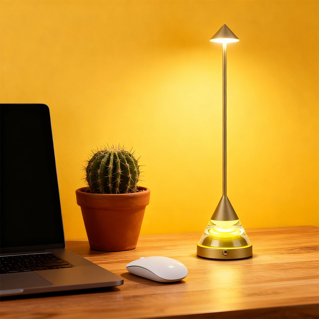A modern gold unique small table lamp illuminating a desk with a laptop and cactus, against a vibrant yellow wall.
