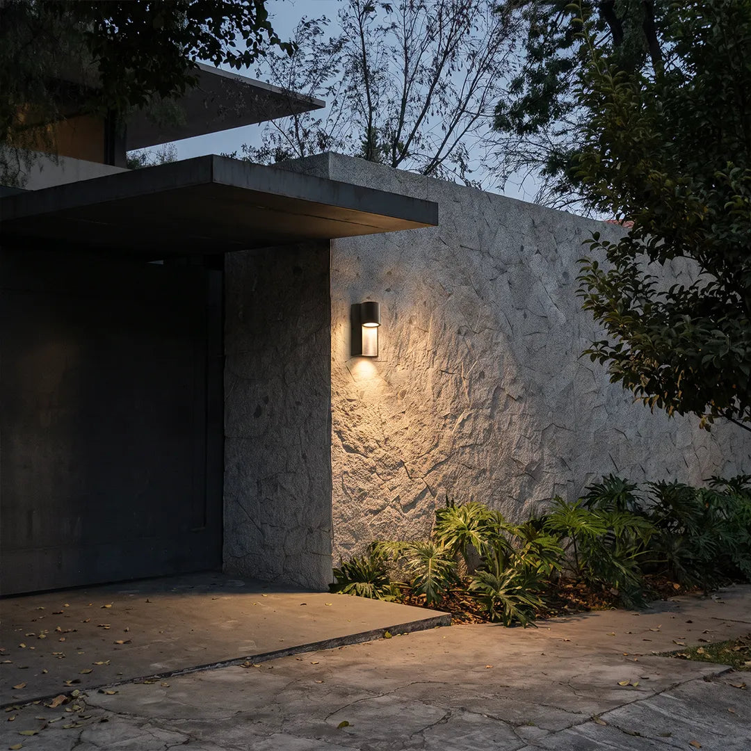 Modern LED outdoor wall light illuminating a textured exterior wall beside a landscaped walkway.