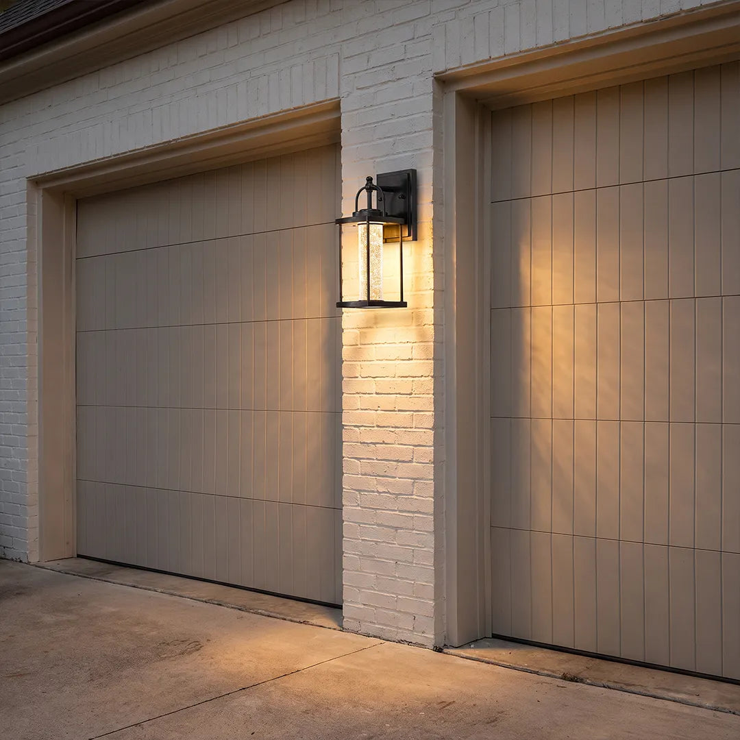 Modern outdoor lantern wall sconce mounted beside a garage door on a white brick exterior.