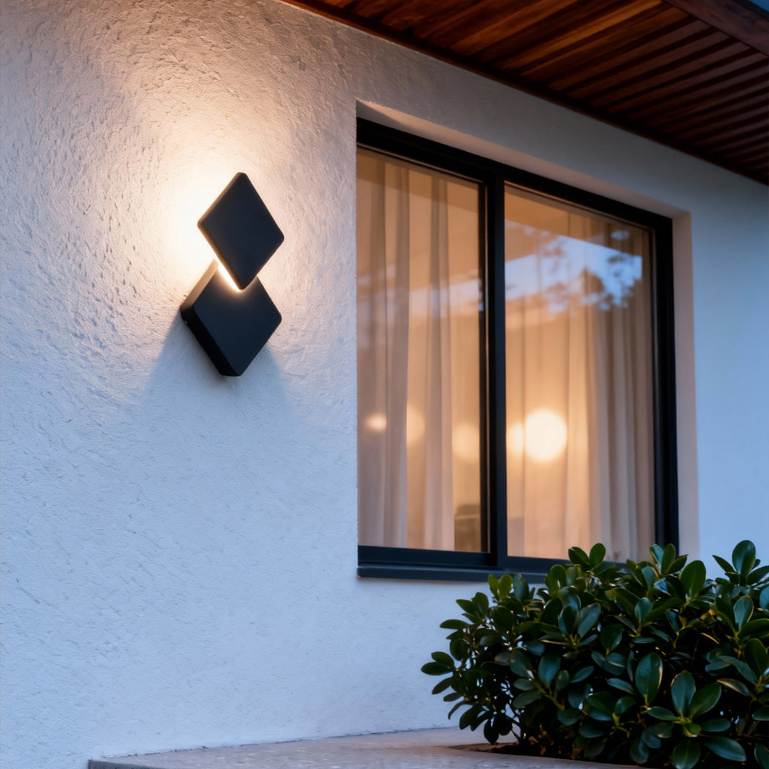 Warm glowing outdoor lighting sconce mounted on a white exterior wall, illuminating the window area with a modern geometric design.