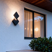 Warm glowing outdoor lighting sconce mounted on a white exterior wall, illuminating the window area with a modern geometric design.
