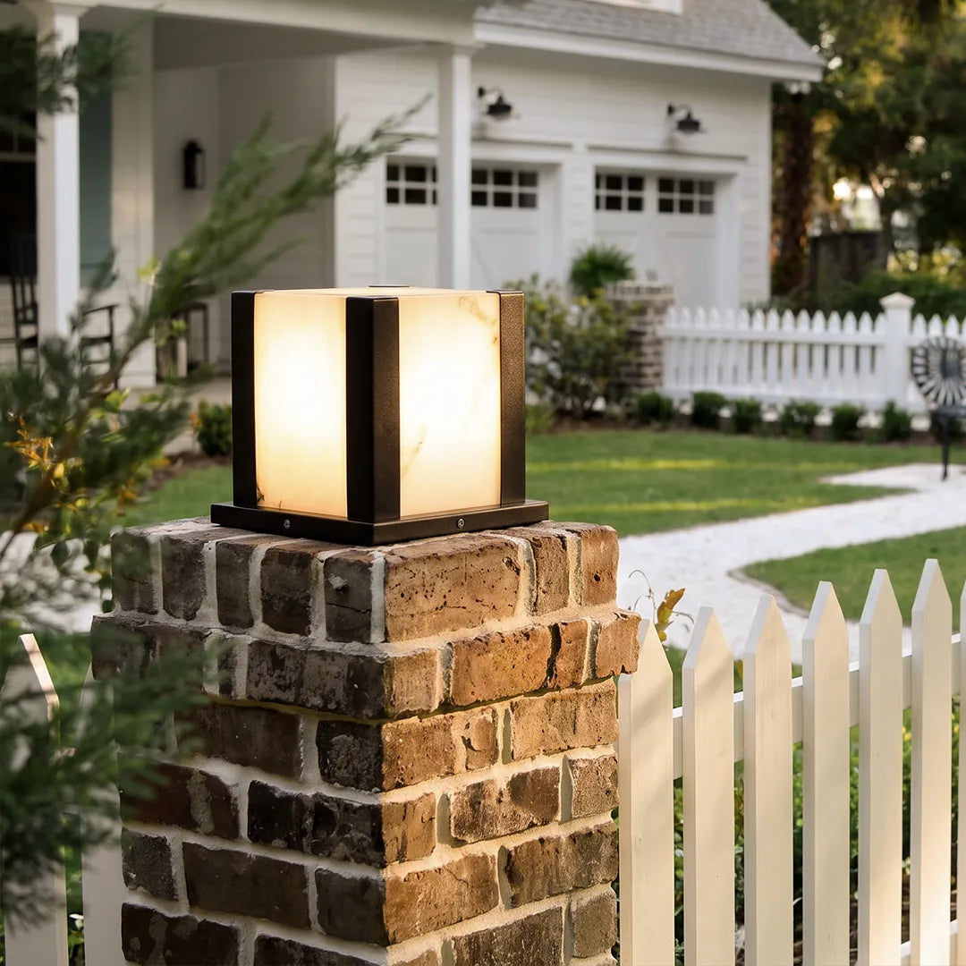 Modern outdoor pillar light with warm glow on brick column beside white picket fence