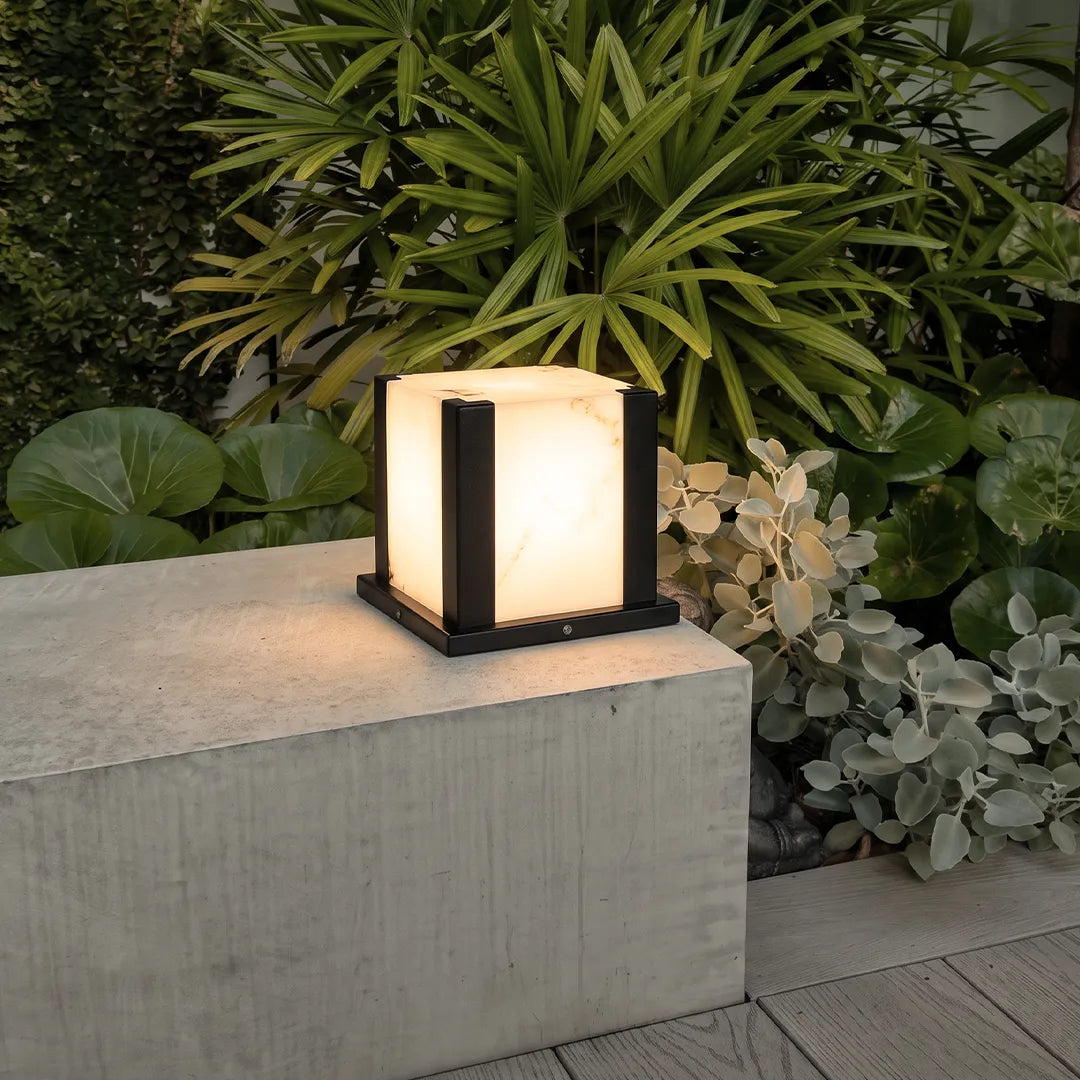 Modern outdoor pillar light with warm glow on concrete wall beside tropical plants