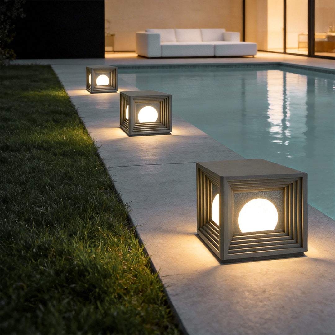Modern outdoor pillar lights with a geometric cube design, illuminating a poolside pathway.