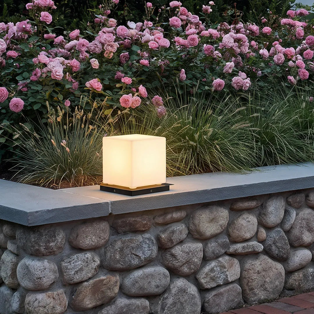 Modern outdoor pillar lights installed on a stone garden wall, adding soft illumination beside blooming pink flowers.