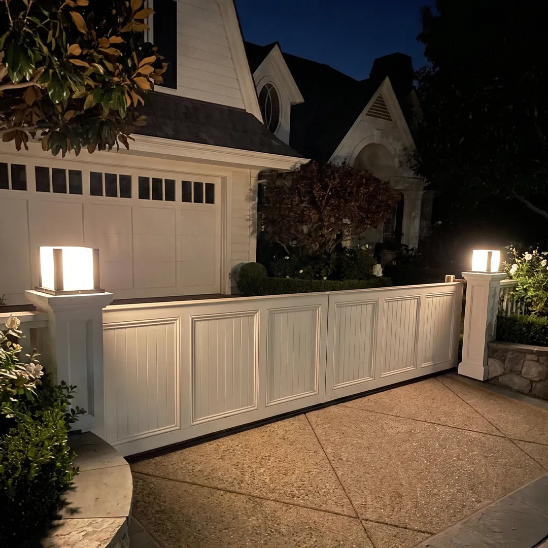 Modern outdoor pillar lights with warm glow along white fence beside driveway