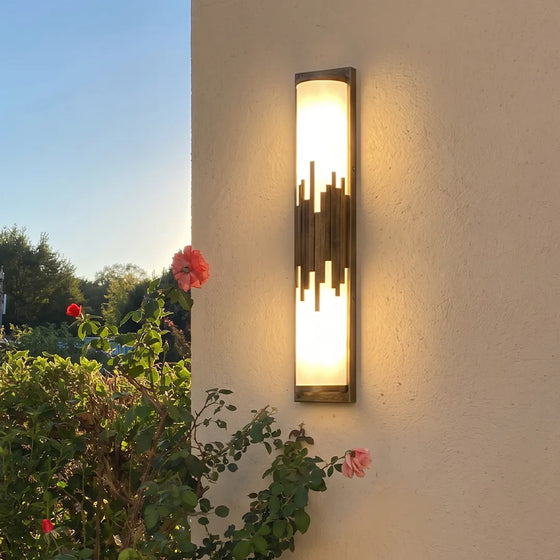 Modern outdoor wall sconce with black and gold accents beside garden flowers