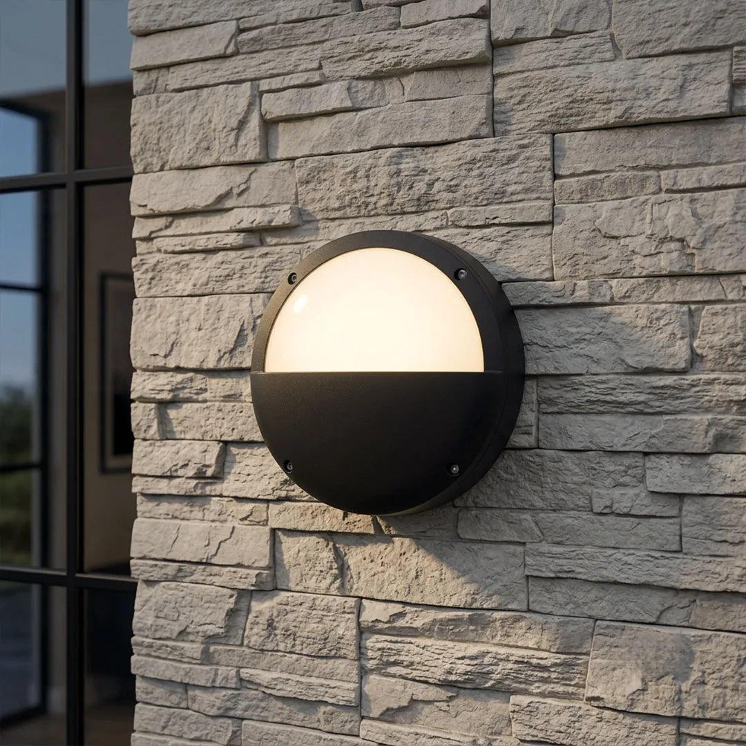 Modern round outdoor wall light fixture with black frame installed on a gray stacked stone exterior wall.