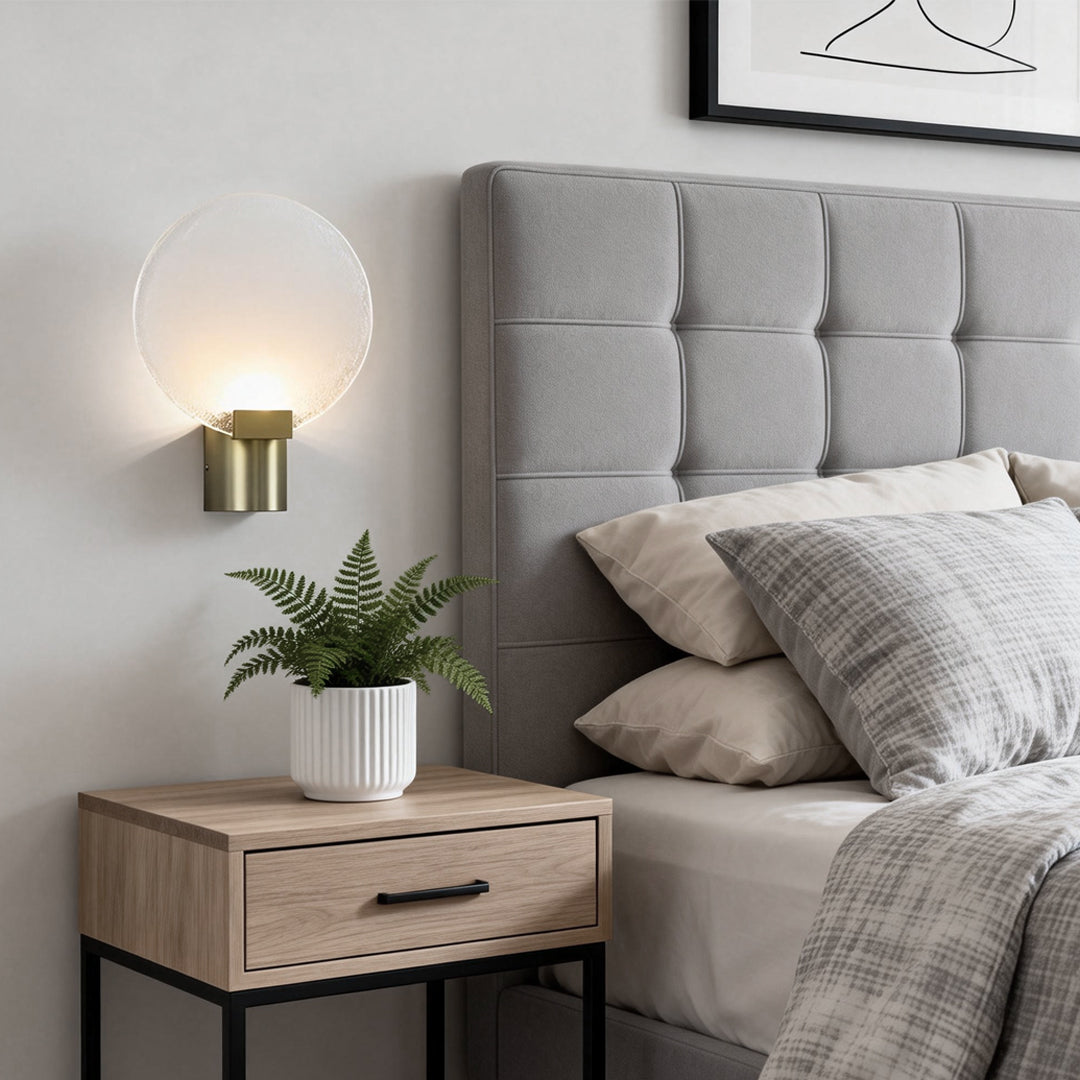 Modern round wall light with a frosted glass shade and gold metal base, mounted beside a cozy bedroom headboard.