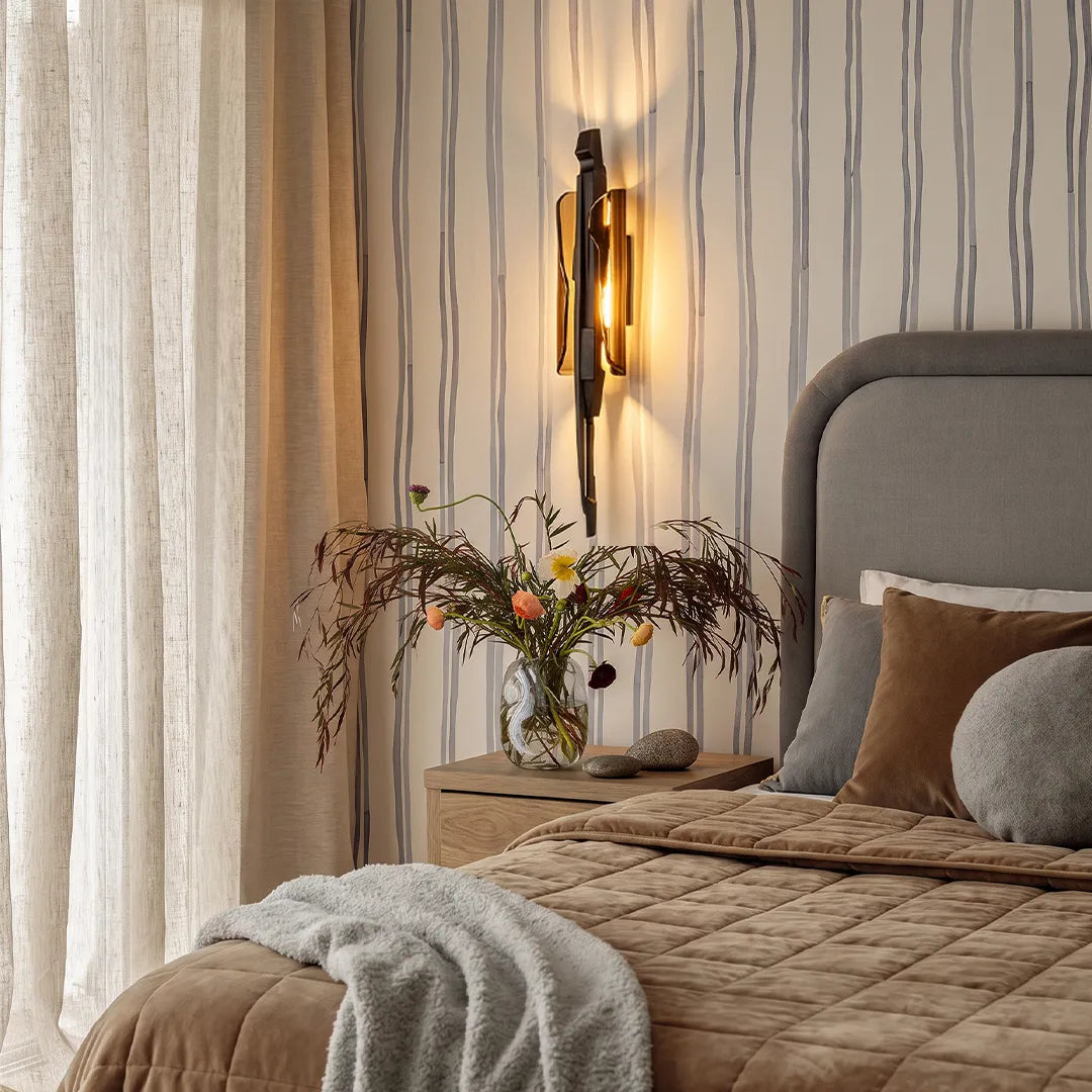 Modern sculptural wall sconce installed beside a bed with soft neutral bedding and striped wall paneling.