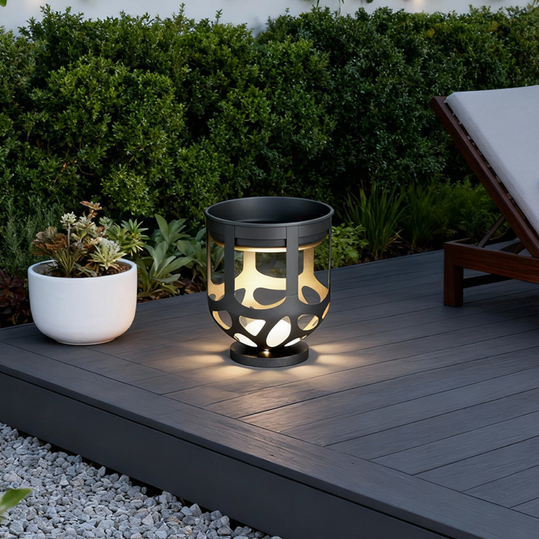 A modern short black lighted outdoor planter with cutout designs glowing on a grey composite deck.