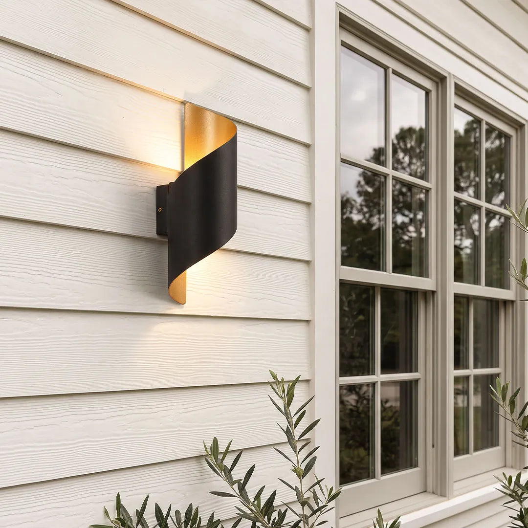 Modern spiral wall light mounted on white exterior siding, featuring a sculptural black and gold curved design with a warm glow.