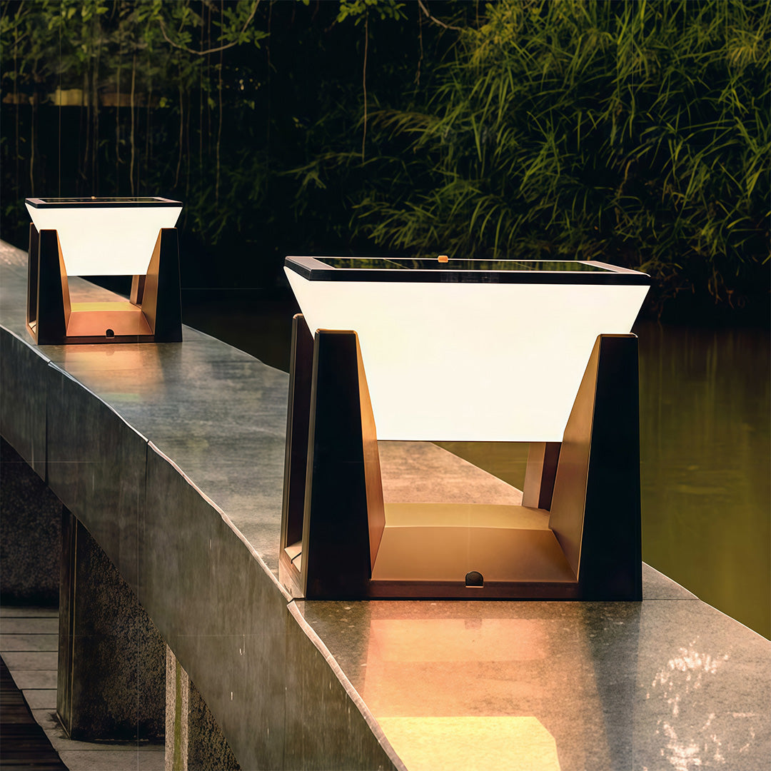 Modern square solar bollard light fixtures glowing warmly on a concrete railing next to a pond at dusk.