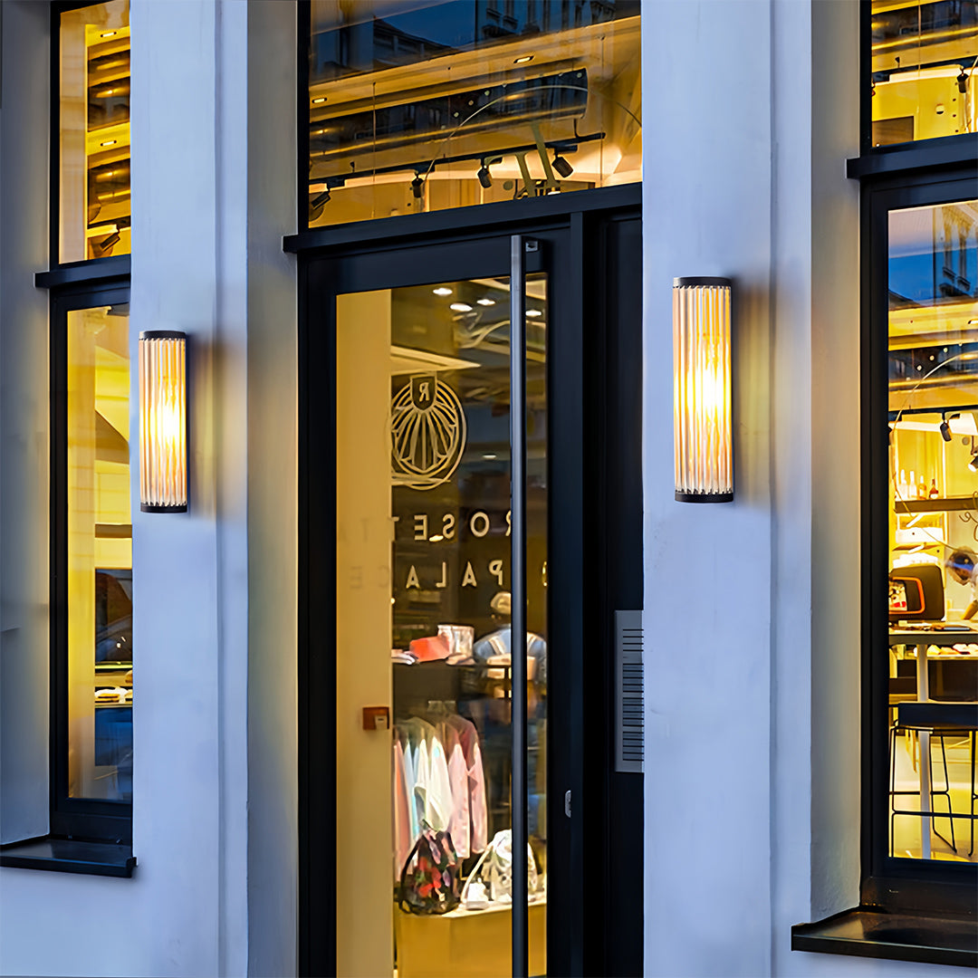 Modern vertical outdoor wall LED light with clear, fluted glass tubes, mounted beside a storefront entrance for commercial appeal.