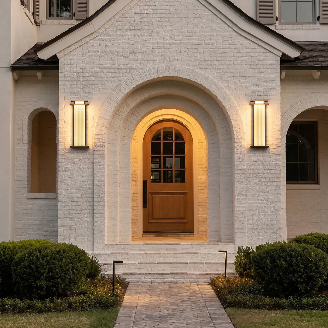 Modern vertical wall sconces installed on both sides of an arched front door on a white brick facade.
