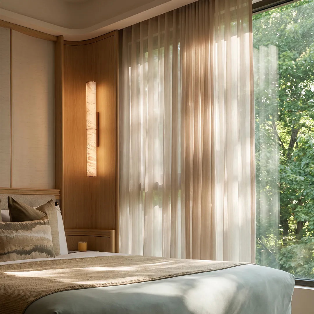 Modern vertical wall sconce illuminating a bedroom corner beside tall curtains and a wood-paneled wall.