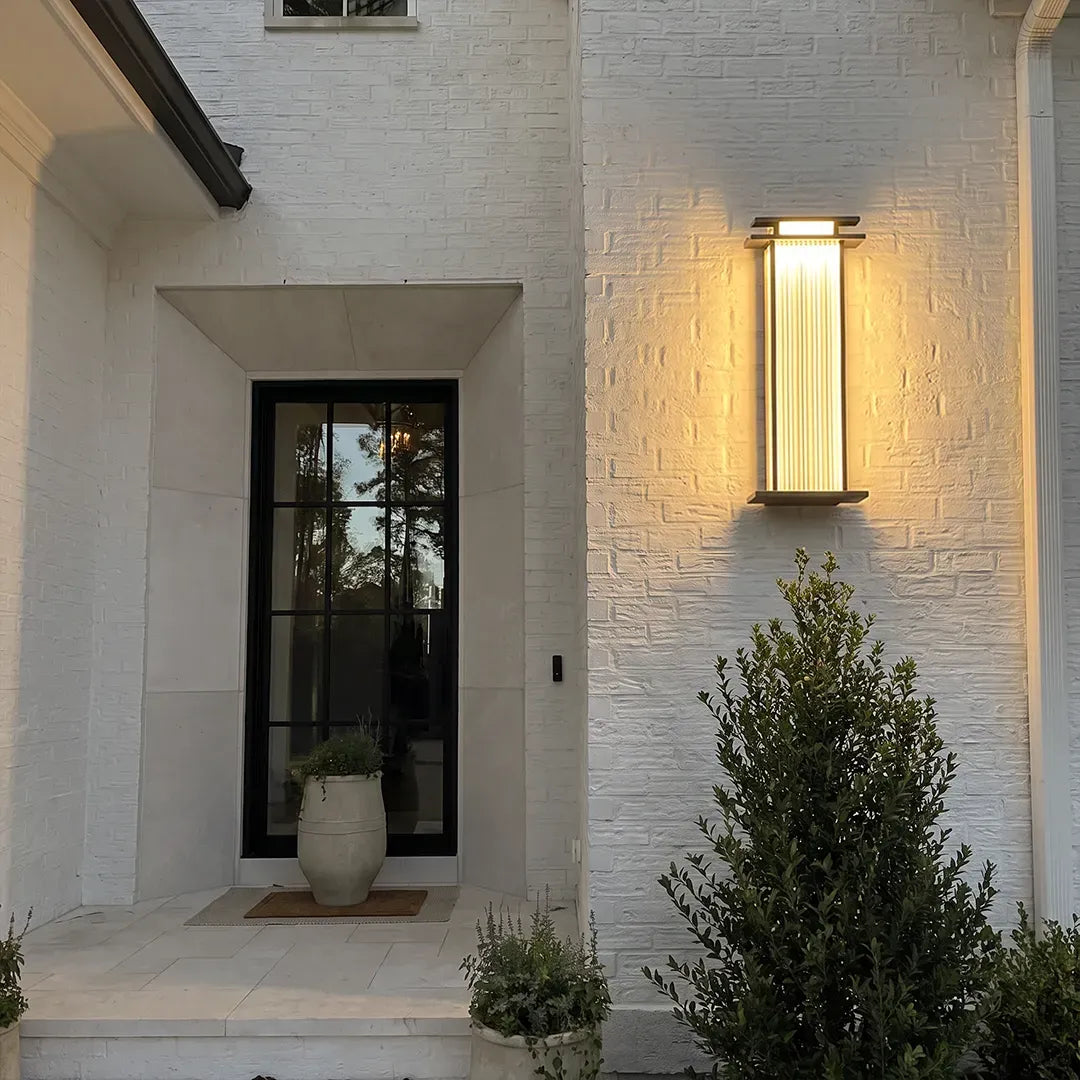 Modern vertical wall sconce mounted beside a front door on a white brick exterior wall.