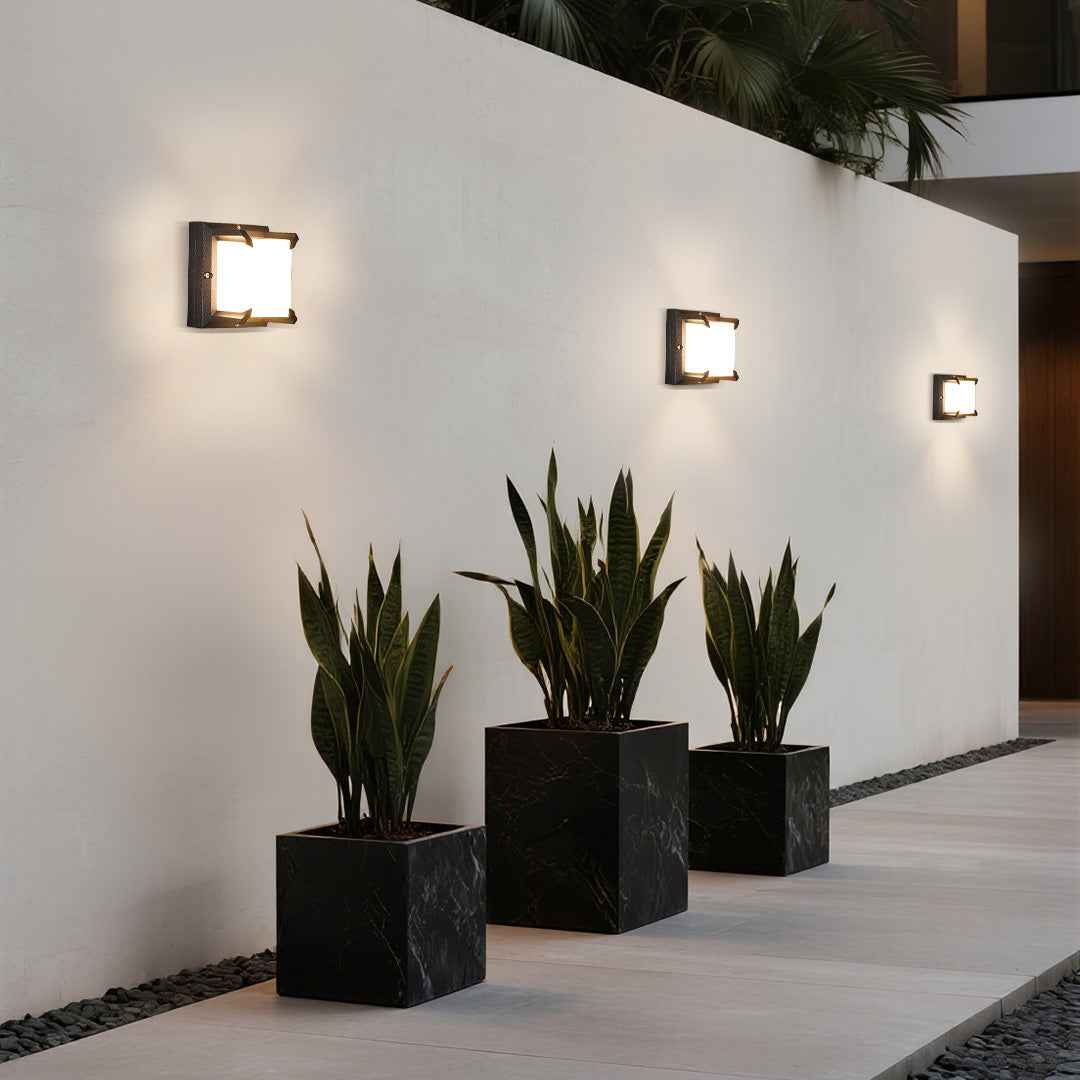Modern walkway illuminated by three sequential square Black Outdoor Wall Light fixtures on a smooth white privacy wall.