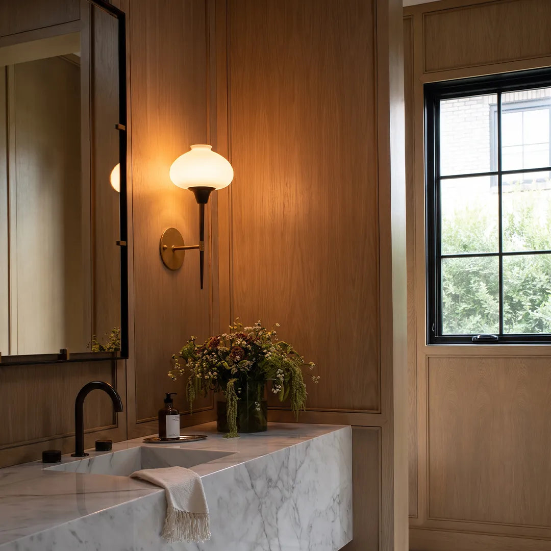 Modern wall light fixture mounted beside a bathroom mirror with a brass backplate and glass shade.