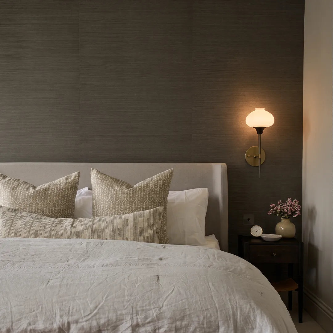 Modern wall light fixture mounted above a bedside table with a glowing glass shade and brass backplate.